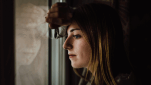 Frau schaut nachdenklich aus dem Fenster, ihr Spiegelbild in der Scheibe – Sinnbild für innere Zerrissenheit und Selbstverleugnung.