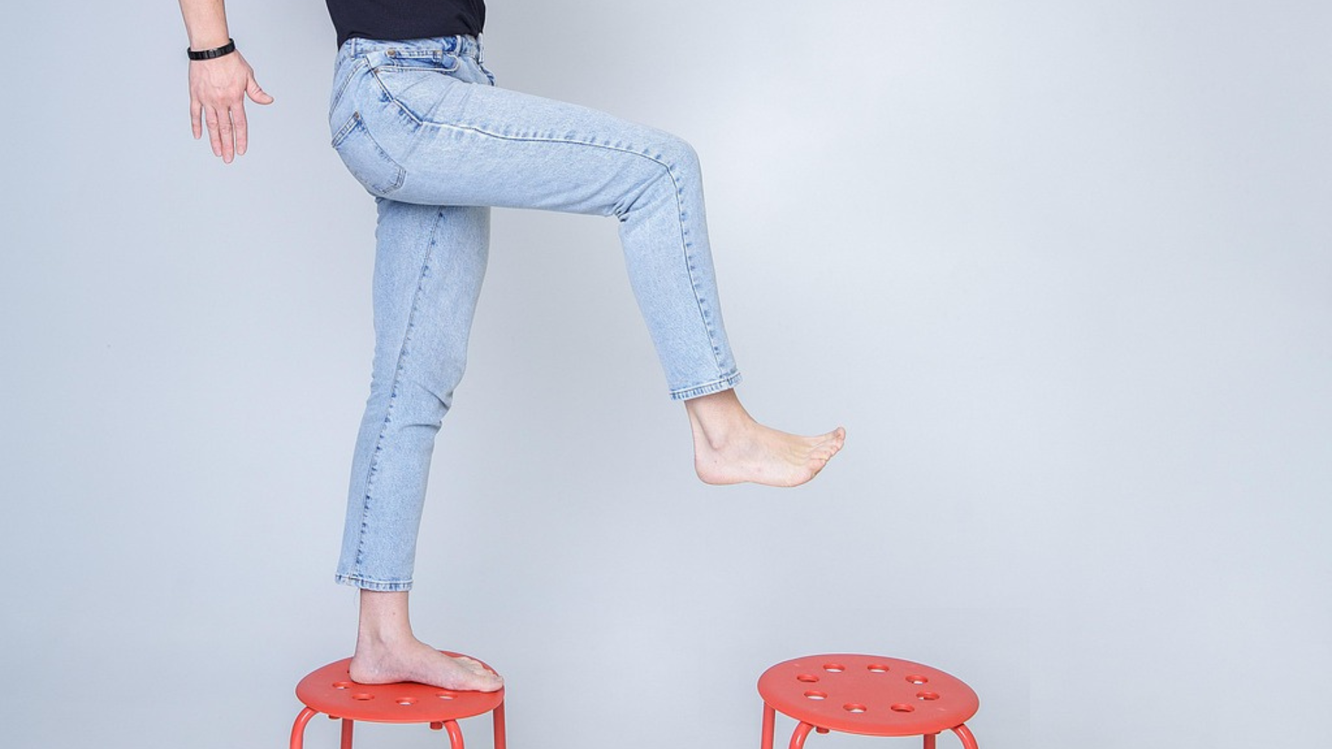 Dein nächster Schritt: zwei Beine - Jeanshose - nackte Füße - Großer Schritt von einem roten Stuhl auf einen zweiten roten Stuhl