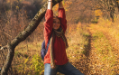 Frau mit rotem Hemd, Jeans und Rucksack häng an einem dicken Ast in herbstlichem Wald und Wiesen Ambiente
