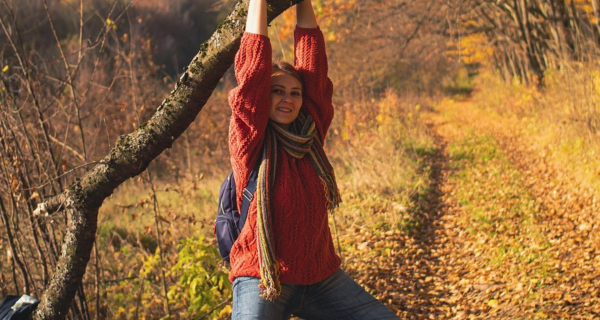 Frau mit rotem Hemd, Jeans und Rucksack häng an einem dicken Ast in herbstlichem Wald und Wiesen Ambiente